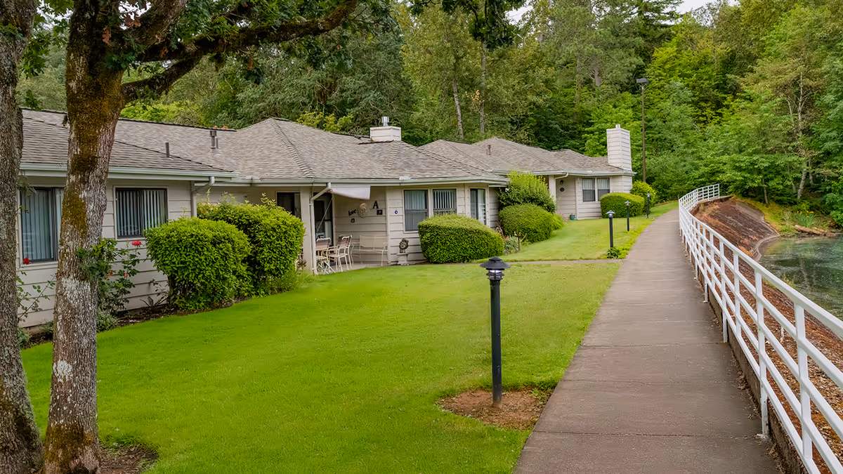 Single-story residential building with a green lawn and a paved riverside walkway with white railing.