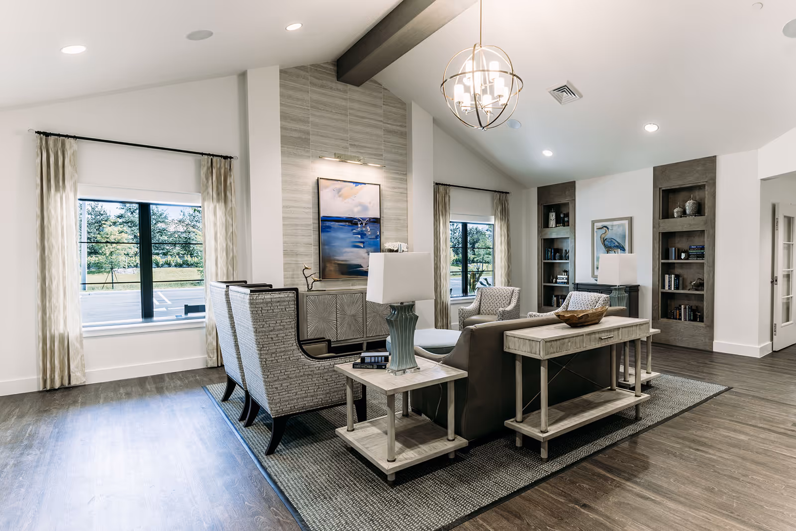 A bright and spacious senior living facility common area with modern furniture including a sofa, armchairs, side tables with lamps, and built-in bookshelves. Large windows with curtains let in natural light, and a decorative painting hangs above a console table against a tiled accent wall.