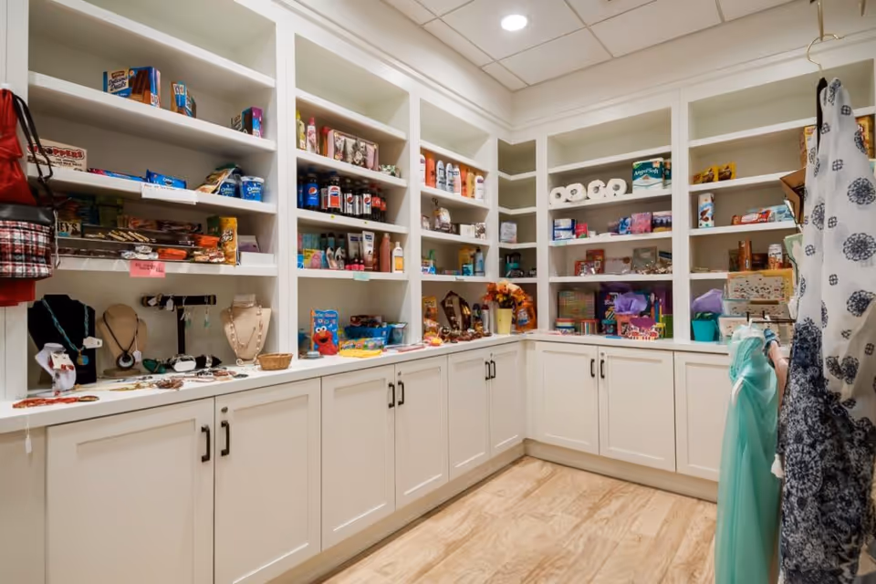A well-lit room with white built-in shelves and cabinets filled with various items including snacks, beverages, toiletries, jewelry, and small gift items. There are also some hanging clothes visible on the right side.