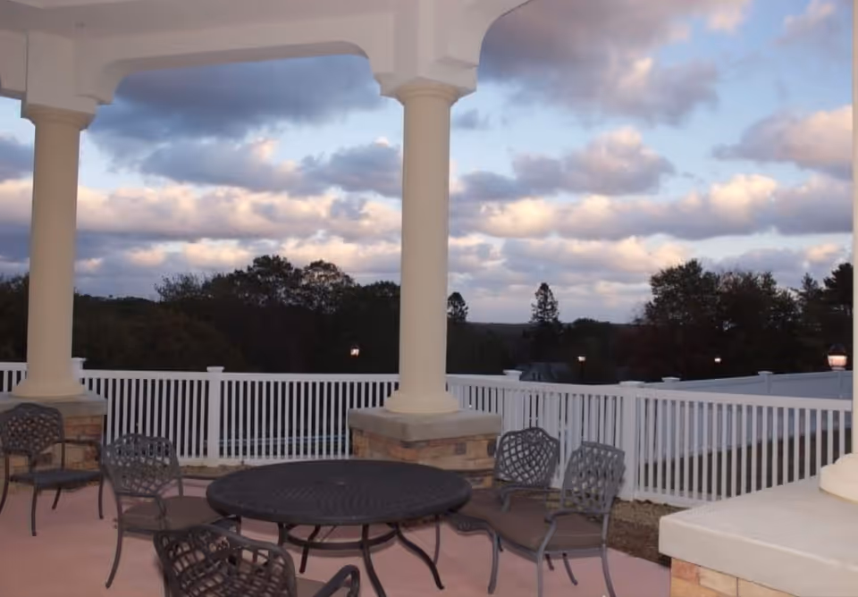 Outdoor patio area with a round metal table and several metal chairs with cushions, surrounded by white fencing and supported by large white columns. Trees and a cloudy sky are visible in the background.