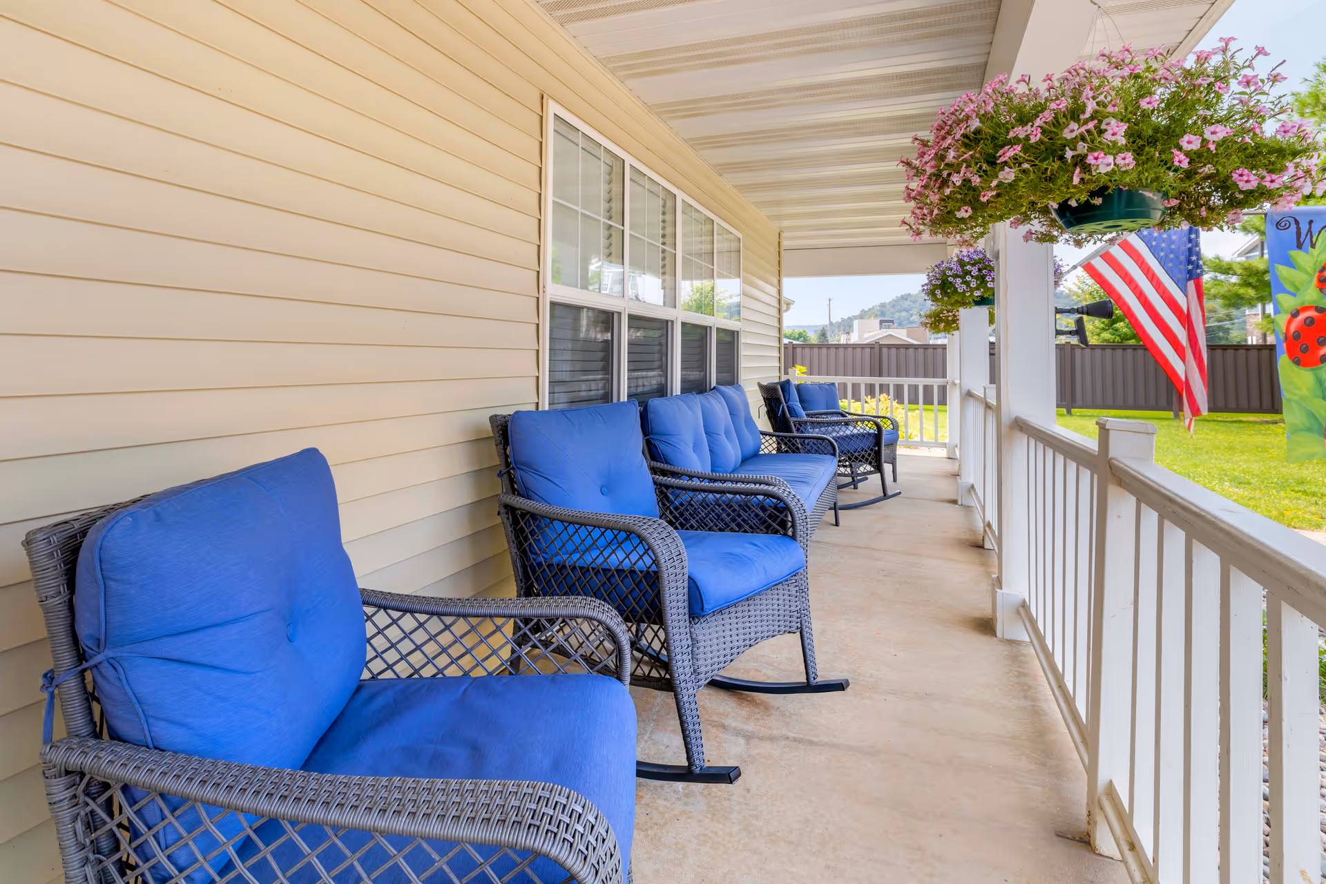 A covered porch with several black wicker rocking chairs featuring blue cushions lined up against the beige siding of a building. Hanging flower baskets with pink and purple flowers are suspended from the porch ceiling. An American flag and a colorful garden flag are visible near the white railing, with a green lawn and a fence in the background.