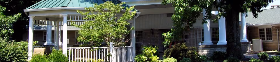 Exterior view of a senior living facility with a covered porch supported by white columns, surrounded by green trees and bushes under a clear sky.
