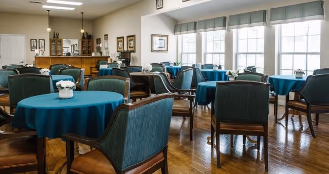 A bright dining room with multiple round tables covered with teal tablecloths, each table adorned with a small white flower centerpiece. The room has large windows letting in natural light, wooden flooring, and green upholstered chairs around the tables. The back of the room features a wooden cabinet and framed pictures on the walls.