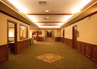 Wide hallway in a senior living facility with beige walls, wooden paneling, and green carpet featuring a decorative square pattern in the center. The hallway is well-lit with ceiling lights and has multiple doorways and windows along the sides.