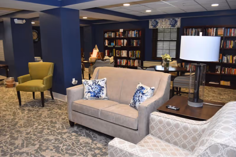 Cozy communal lounge with a beige loveseat, patterned armchairs, a green accent chair, side table with lamp, and bookshelves along a navy-blue wall.