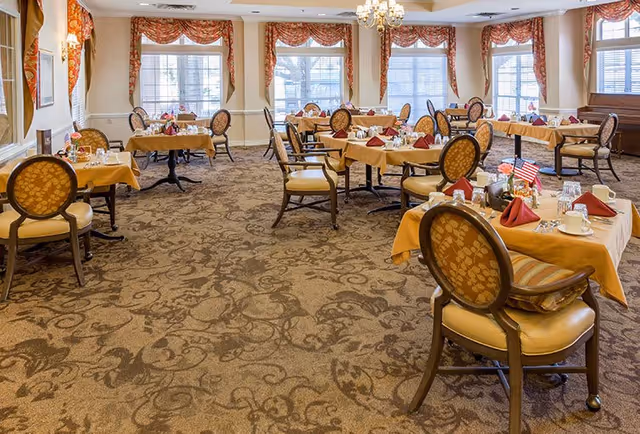 A spacious dining room with multiple tables covered in yellow tablecloths, each set with red napkins, glassware, and silverware. The room features large windows with decorative curtains, patterned carpet, and elegant chairs with cushioned seats and backs. A chandelier hangs from the ceiling, adding a touch of sophistication.