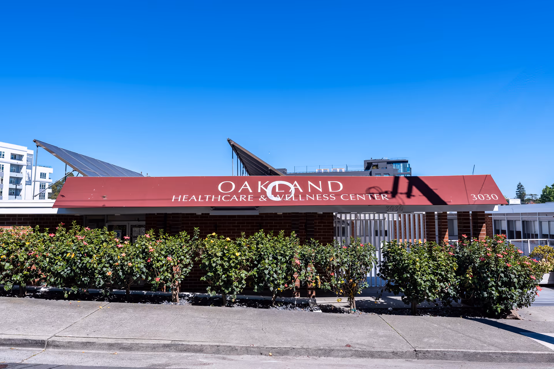 Front exterior of Oakland Healthcare & Wellness Center with a maroon awning, shrubs, and clear blue sky.