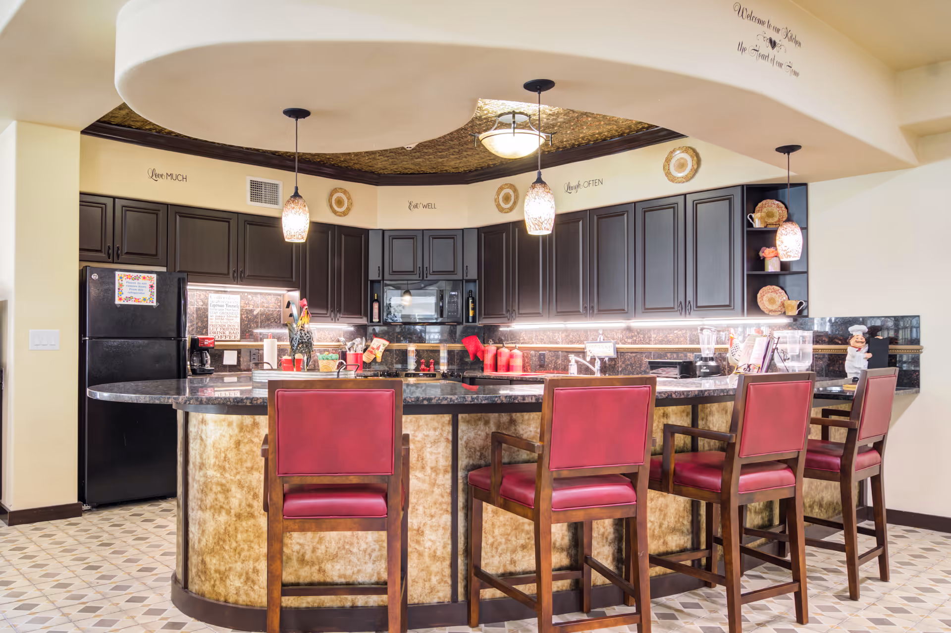 A well-lit kitchen area with a curved island countertop surrounded by four wooden chairs with red cushions. The kitchen features dark cabinetry, a black refrigerator, a microwave, and various kitchen items such as a coffee maker, blender, and decorative pieces. Pendant lights hang from the ceiling, and the floor has a patterned tile design.