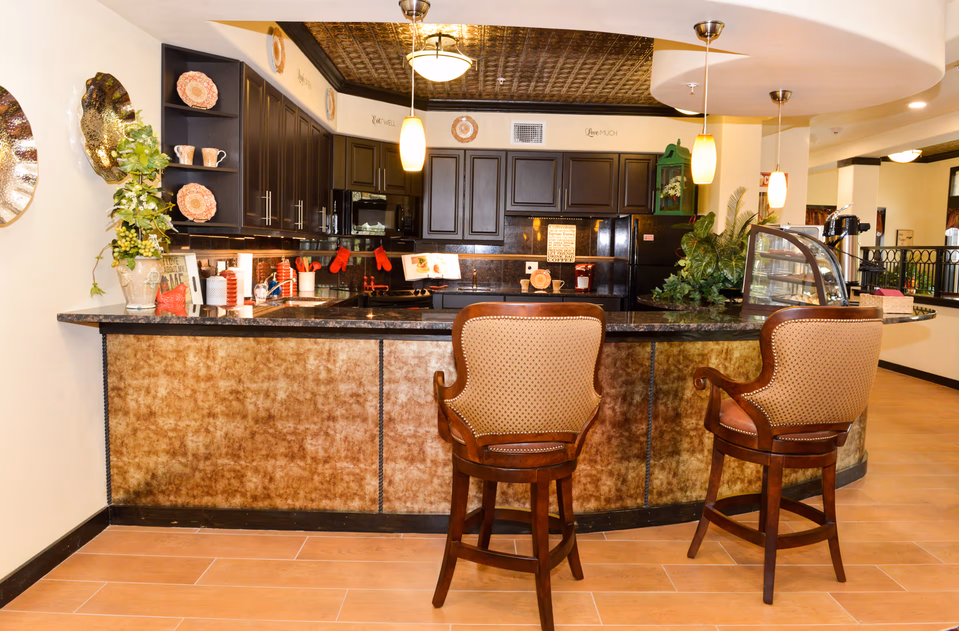 Interior view of a kitchen area in a senior living facility with a curved counter, two upholstered bar stools, dark wood cabinets, hanging pendant lights, and decorative plants and plates.