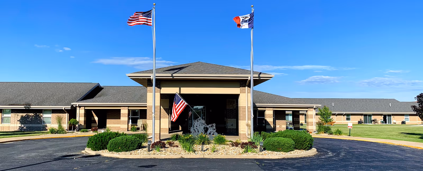 Front exterior view of River Bend Retirement Community building with two flagpoles displaying the American flag and another flag. The building is single-story with a covered entrance and landscaped greenery in front. The sky is clear and blue.