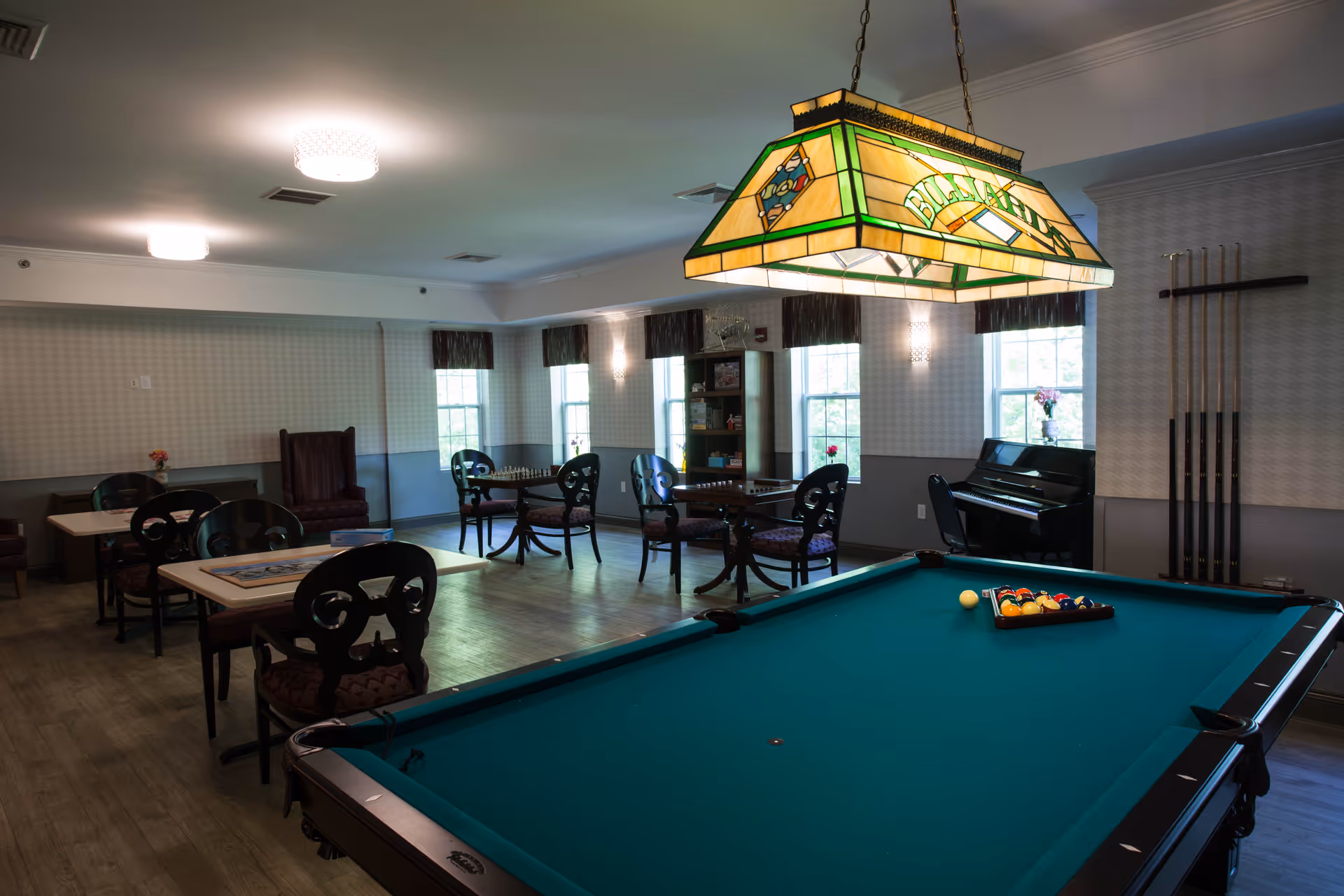 A recreational room with a pool table in the foreground, a stained glass billiards light hanging above it, several tables and chairs arranged around the room, a piano in the corner, and windows letting in natural light.