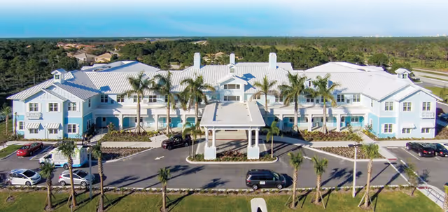 Front exterior of a two-story light-blue senior living building with a covered porte-cochère, palm trees, and cars in the driveway.