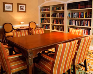 A cozy interior room featuring a large wooden table surrounded by six striped upholstered chairs. In the background, there are built-in bookshelves filled with books and two framed pictures on the wall above a small table with two wooden chairs and a lamp.