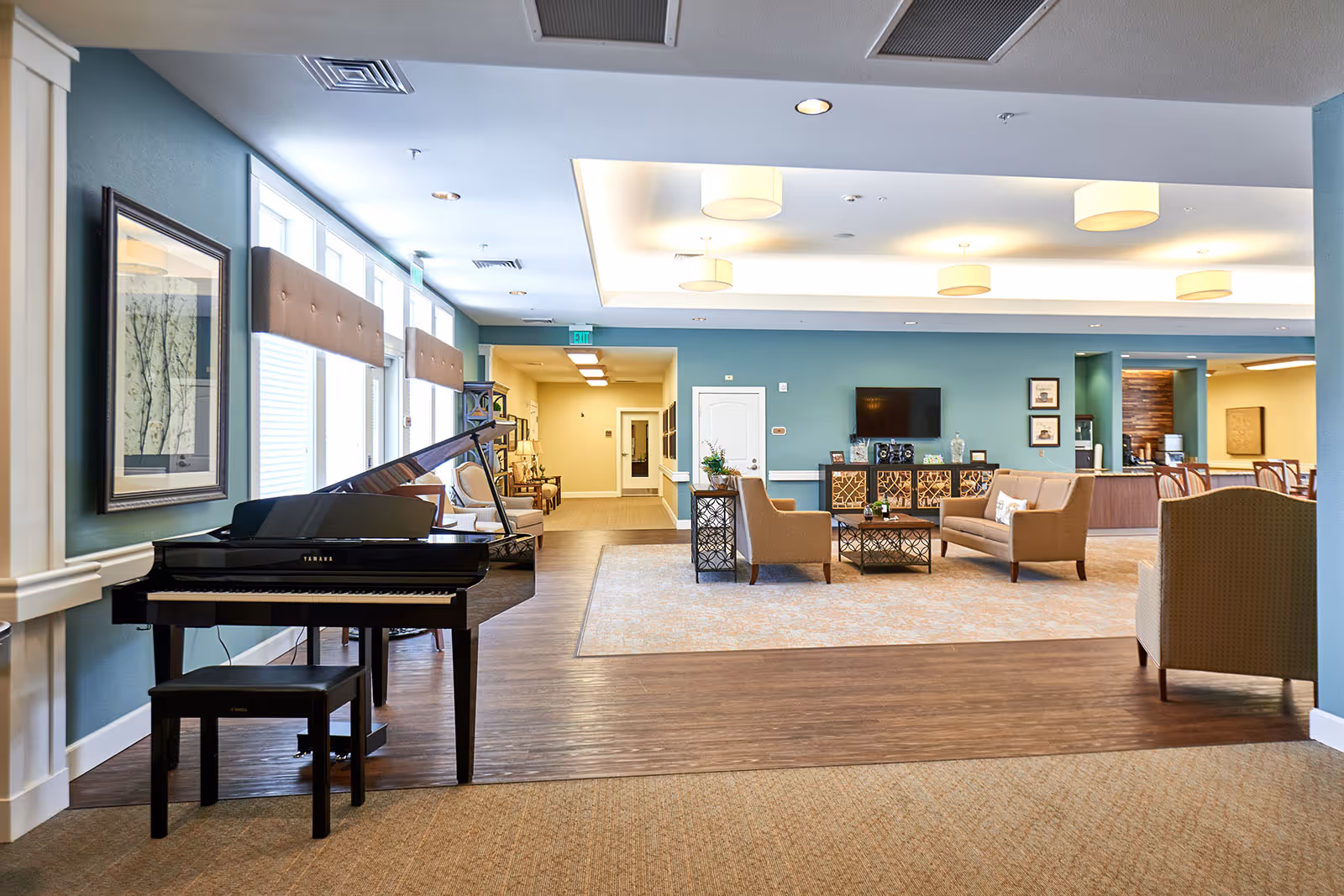 A spacious and well-lit common area in a senior living facility featuring a black Yamaha grand piano with a bench, several beige armchairs and sofas arranged around a coffee table on a large area rug, a flat-screen TV mounted on a teal accent wall, decorative framed pictures, and a hallway leading to other rooms.