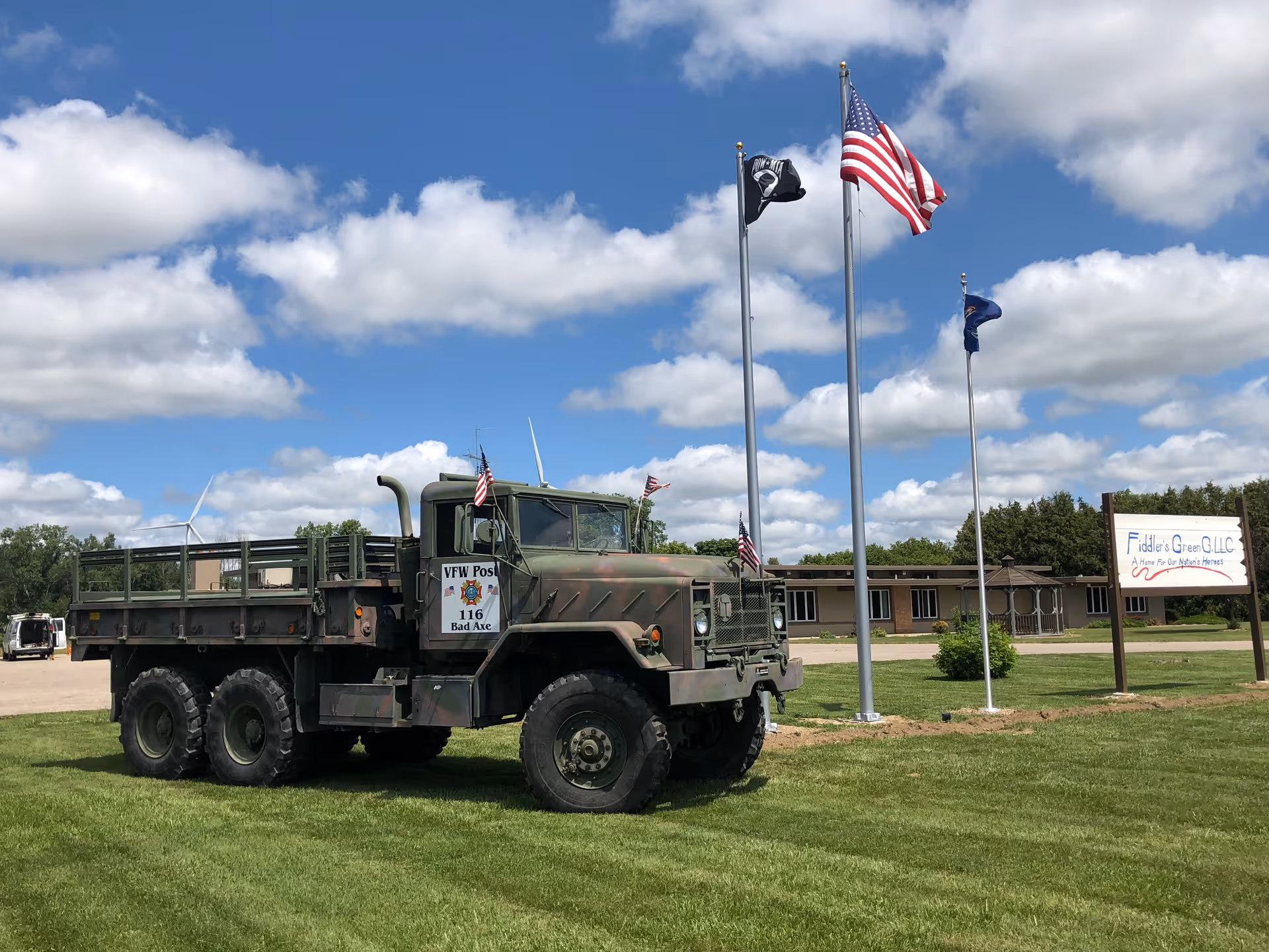 A military-style truck with camouflage paint is parked on a grassy area near three flagpoles flying the American flag, a POW/MIA flag, and another flag. In the background, there is a single-story building and a sign that reads 'Fiddler's Green G.L.L.C. A Home For Our Nation's Heroes' under a partly cloudy blue sky.