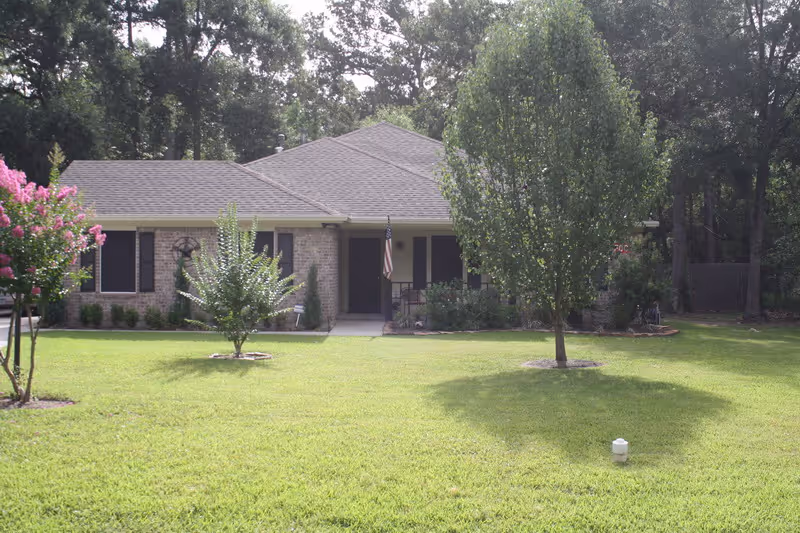 Front exterior view of a single-story brick house with a gray roof, black shutters, and a small porch with an American flag. The house is surrounded by a well-maintained lawn with two small trees and some bushes, and there are tall trees in the background.