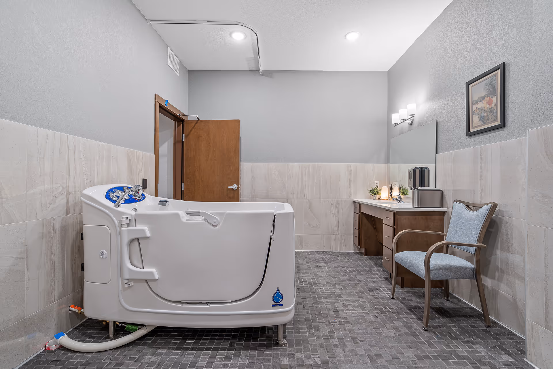 A spacious bathroom featuring a white walk-in bathtub with safety handles and controls, a wooden door, a vanity with a large mirror, a chair with light blue upholstery, and a framed picture on the wall. The walls are tiled halfway up with light beige tiles and painted gray above, and the floor is covered with small dark gray tiles.