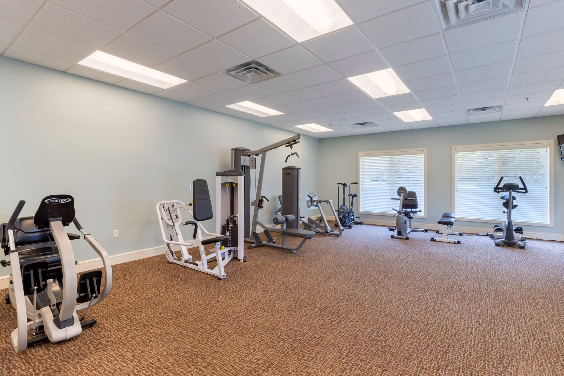 A fitness room with various exercise equipment including a NuStep machine, weight machines, stationary bikes, and other workout apparatus. The room has light blue walls, a brown carpeted floor, and large windows with blinds allowing natural light to enter.