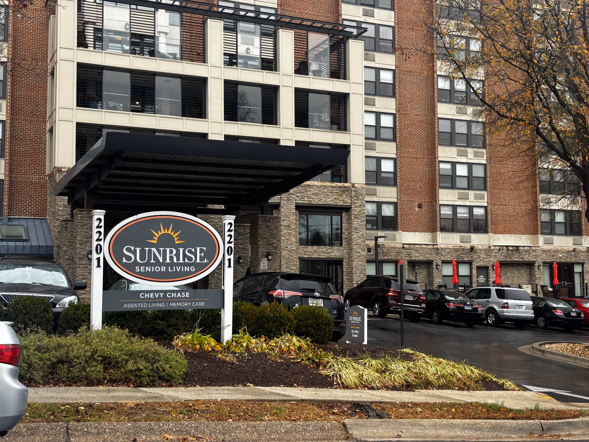 Front entrance of the Sunrise of Chevy Chase senior living building with a large 'Sunrise Senior Living' sign, canopy, and parked cars.