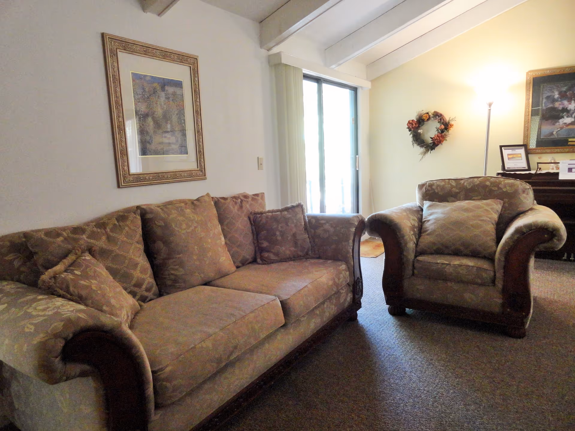 A cozy living room with a patterned brown sofa and matching armchair. There is a framed painting on the wall above the sofa, a floor lamp, a decorative wreath on the wall, and a piano with framed pictures and certificates on top. A sliding glass door with vertical blinds lets in natural light.