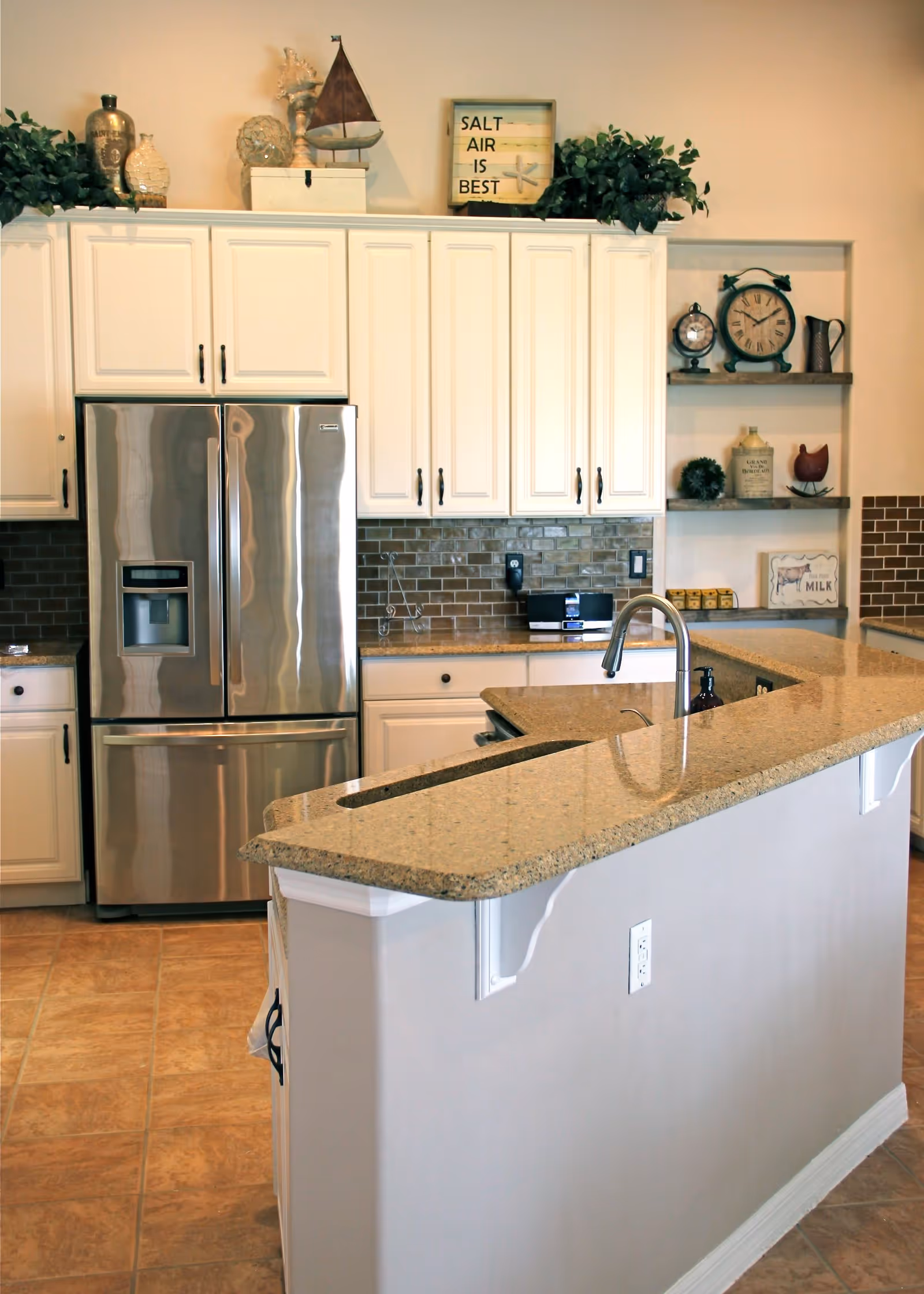 Modern kitchen with white cabinets, a stainless steel refrigerator, a granite countertop island with a built-in sink and faucet, decorative items on top of the cabinets and shelves, and a tiled floor.