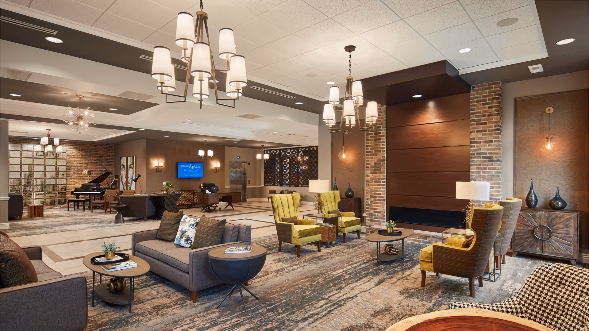 A spacious and elegantly decorated senior living facility common area with modern chandeliers, comfortable seating including sofas and yellow armchairs, a fireplace with brick accents, a grand piano, and a reception desk with a screen displaying Belmont Village Senior Living Lincoln Park.
