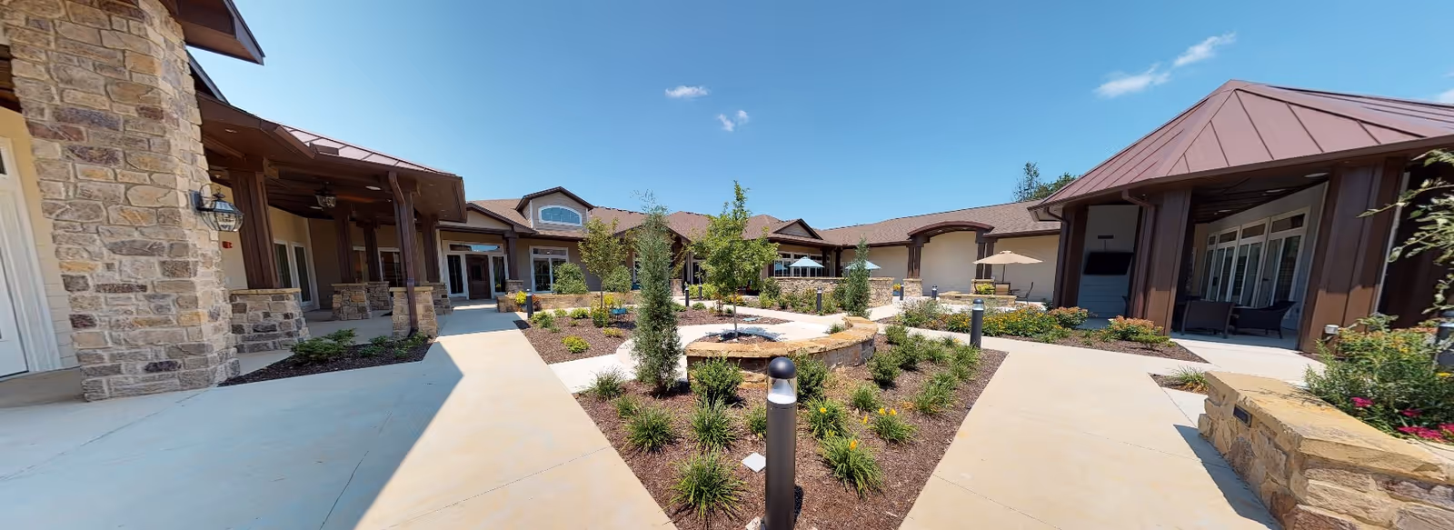 Outdoor courtyard area of Grand Brook Memory Care of Zionsville featuring a landscaped garden with small trees and shrubs, surrounded by a stone and wood building with covered walkways and patio seating under umbrellas on a sunny day with a clear blue sky.