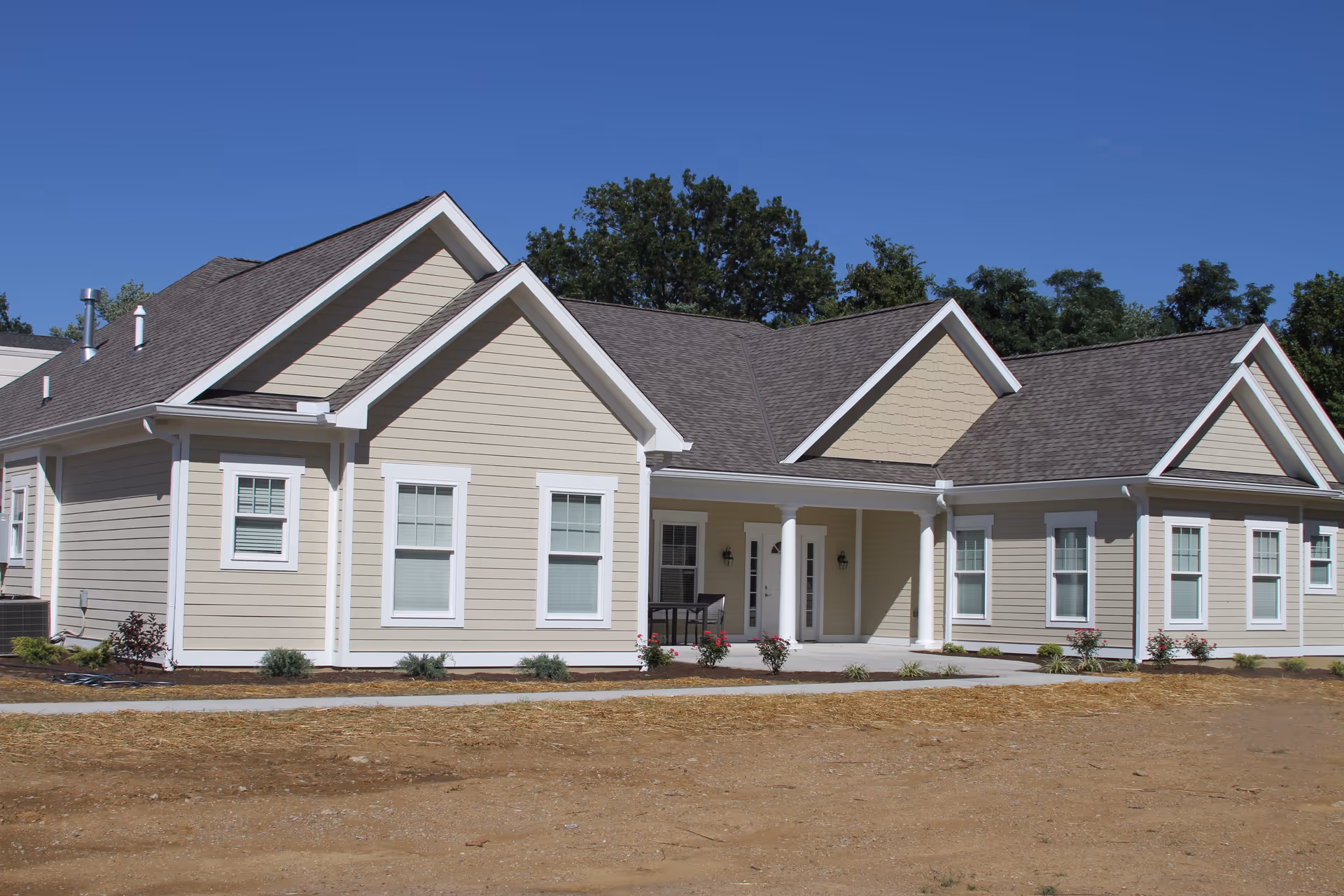 Exterior view of a single-story beige senior living facility building with white trim, multiple windows, a covered entrance with columns, and a clear blue sky in the background.