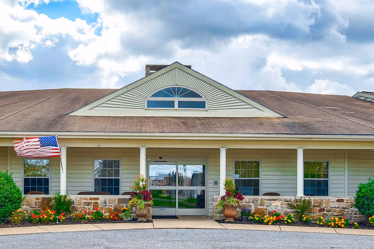 Single-story facility entrance with a covered porch framed by white columns, an American flag, potted plants and flowerbeds under a cloudy sky.