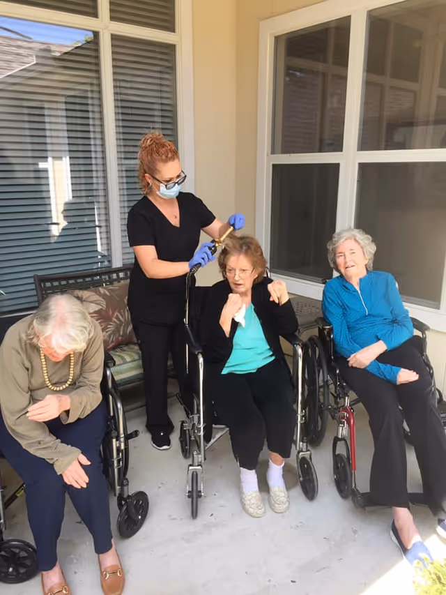 A caregiver wearing a mask and gloves is styling the hair of an elderly woman sitting in a wheelchair on a covered patio. Two other elderly women, also in wheelchairs, are seated nearby, one looking down and the other smiling. The setting appears to be an outdoor area of a care home with windows and a bench in the background.