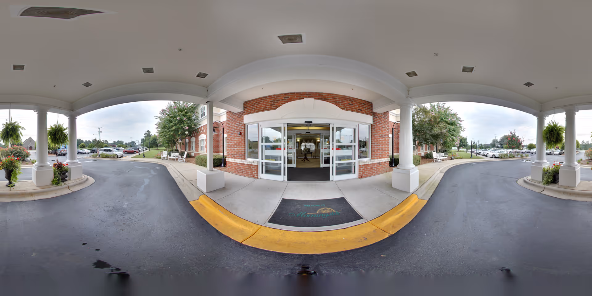 Covered entrance of a senior living facility with automatic sliding glass doors, brick exterior walls, white columns, and a driveway for vehicle drop-off. There are hanging plants and flower pots on either side of the entrance, with benches along the sidewalk. Cars are parked in the parking lot visible in the background under a cloudy sky.
