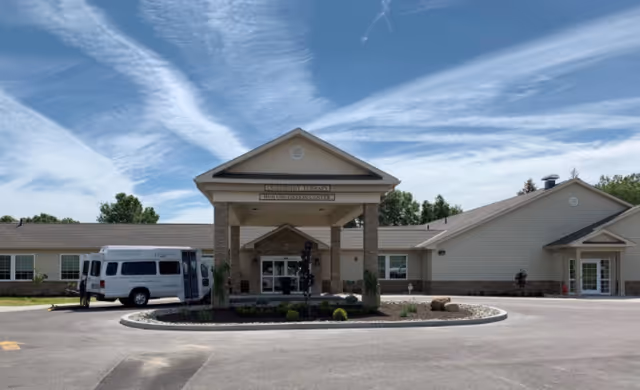 Front exterior view of Woodlands at Austinwoods facility showing a single-story building with a covered entrance, a white van parked on the left side, and a clear blue sky with wispy clouds above.