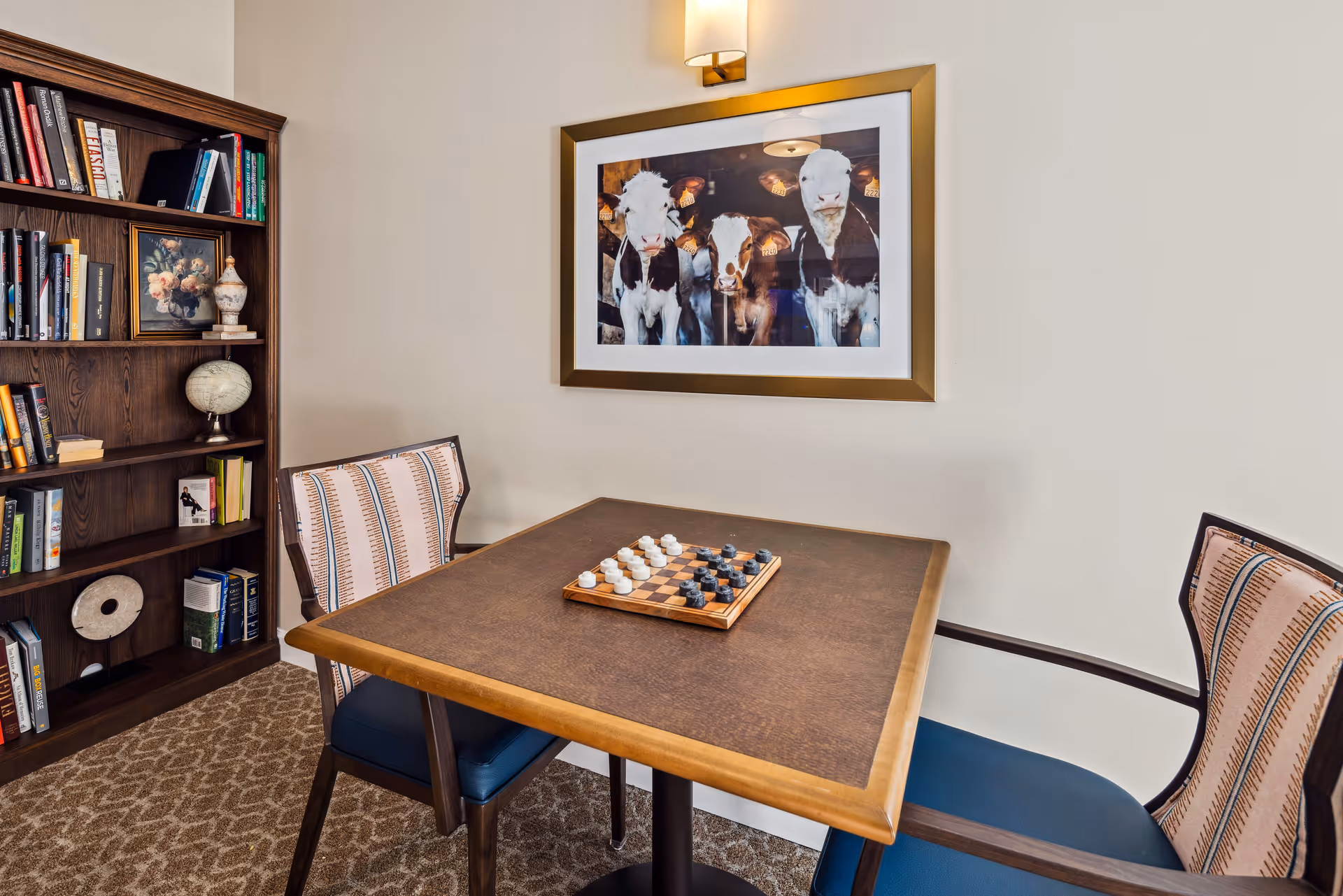 Small seating area with a table set for a board game, two chairs, a bookshelf, and framed cow artwork on the wall.