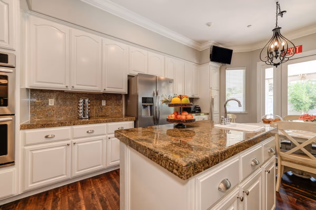 A bright kitchen with white cabinets, granite countertops, a stainless steel refrigerator, a double oven, and a kitchen island with a tiered fruit stand. There is a sink on the island, a chandelier hanging from the ceiling, and a small dining area with chairs near a window and glass door.