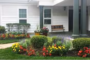 Front exterior of a senior living building with a porch, bench, and colorful flower garden