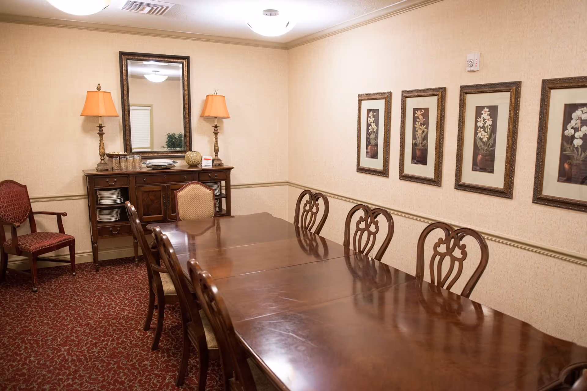 A formal dining room with a long polished wooden table surrounded by wooden chairs with cushioned seats. On the left side, there is a wooden sideboard with plates and decorative items, flanked by two table lamps with orange lampshades. Above the sideboard is a large mirror. The walls are beige with a chair rail and decorated with four framed floral paintings. The floor is covered with a red patterned carpet.