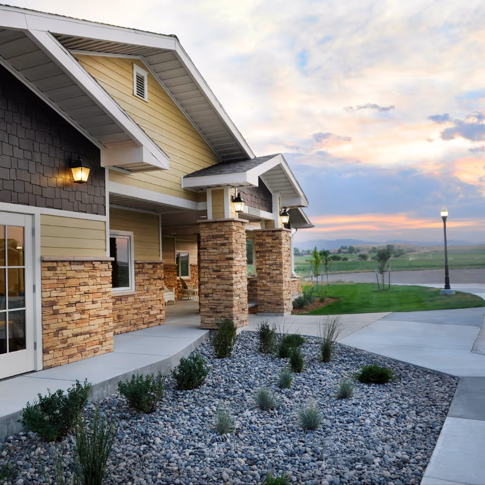 Exterior view of a building at sunset with stone pillars, yellow siding, and a landscaped area with rocks and small bushes in front. A street lamp is visible in the background along a curved driveway.