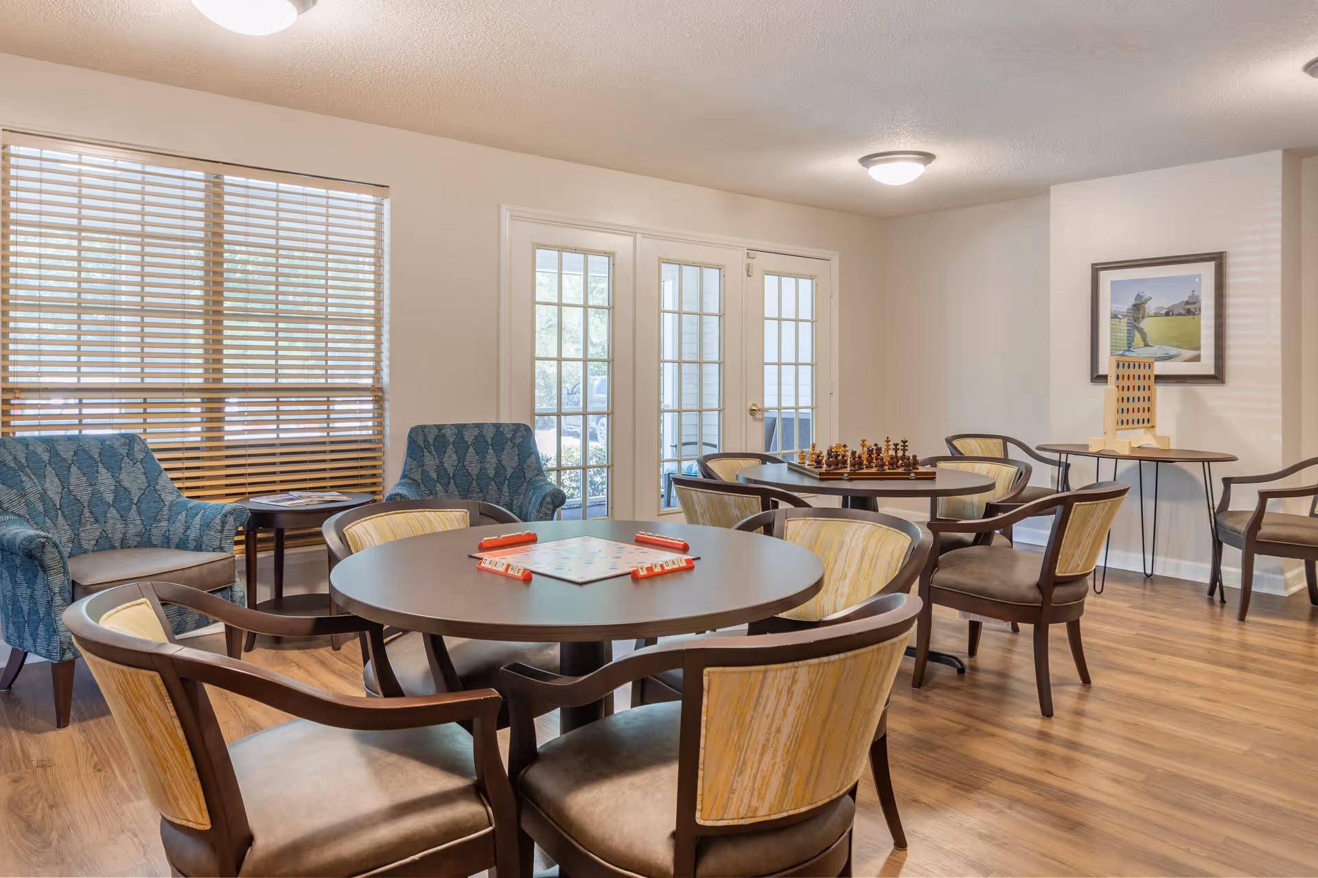 A well-lit common room with wooden flooring featuring two round tables with chairs around them. One table has a Scrabble game set up, and the other has a chessboard. There are two blue patterned armchairs near a window with wooden blinds. A small table with a Connect Four game is against the wall, which also has a framed picture of a golfer. French doors lead outside, allowing natural light into the room.