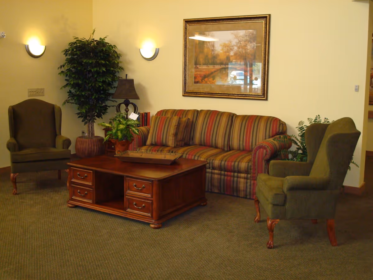 Lounge area with a striped sofa, two green wingback chairs, wooden coffee table, framed wall art, and potted plants.