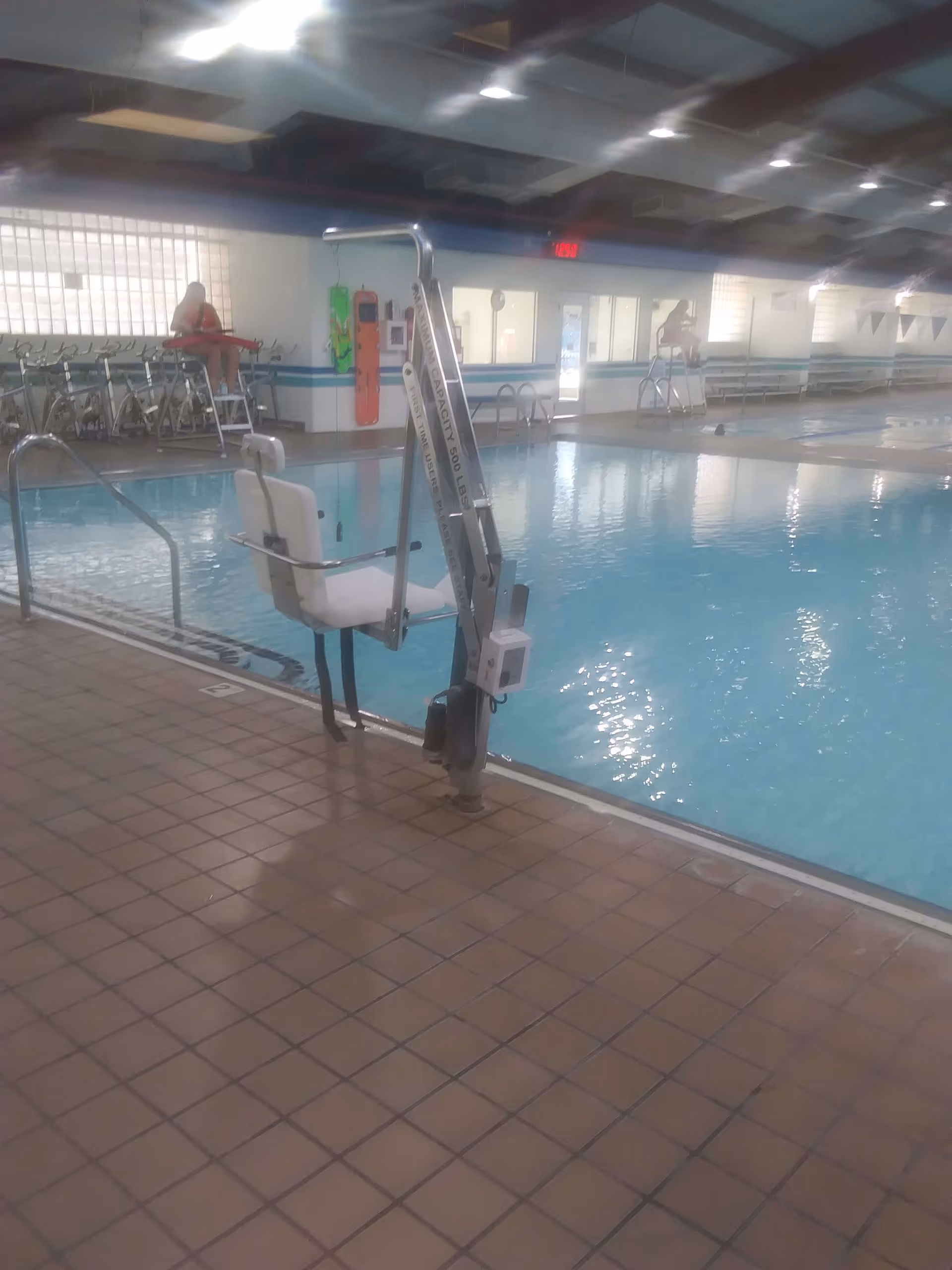 Indoor swimming pool with a pool lift chair for accessibility, tiled floor surrounding the pool, and two lifeguards seated on elevated chairs in the background.