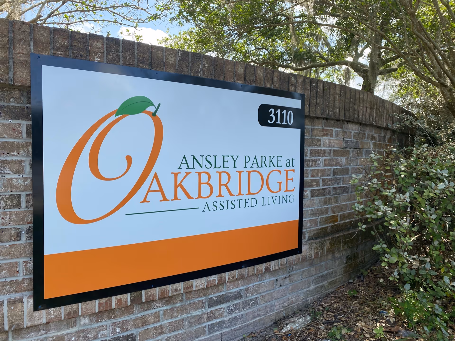 A large sign reading "Ansley Parke at Oakbridge Assisted Living" with the number 3110 mounted on a brick wall surrounded by shrubs.