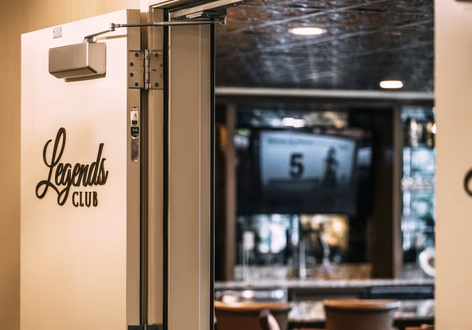 Partially open door with a sign that reads 'Legends Club' leading into a room with a bar counter and stools, and a television screen mounted on the wall in the background.