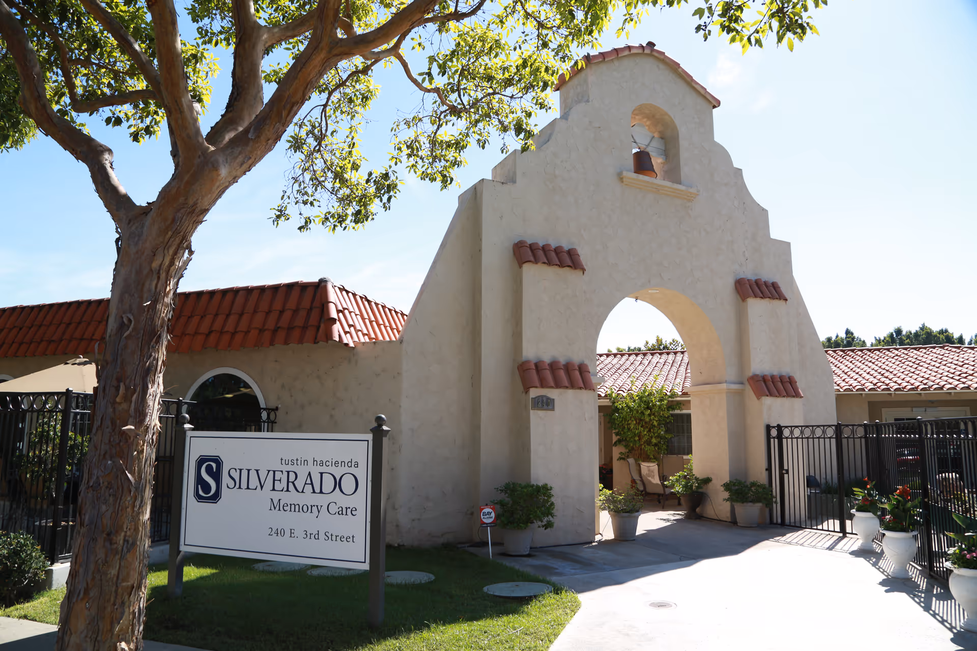 Exterior view of Silverado Tustin Hacienda Memory Care Community featuring a stucco archway with red tile accents, a gated entrance, potted plants, and a sign displaying the facility's name and address at 240 E. 3rd Street.