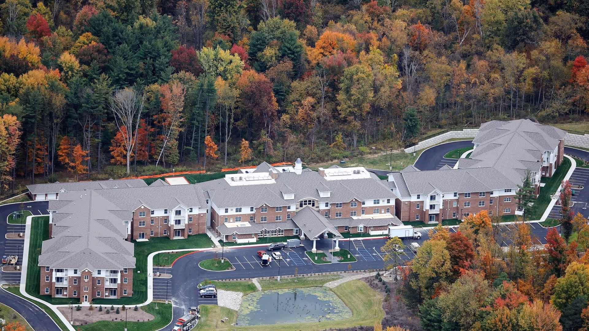 Aerial view of Hudson Meadows Retirement Community surrounded by colorful autumn trees. The large, multi-wing building has a gray roof and brick exterior with a parking lot and a small pond in front.