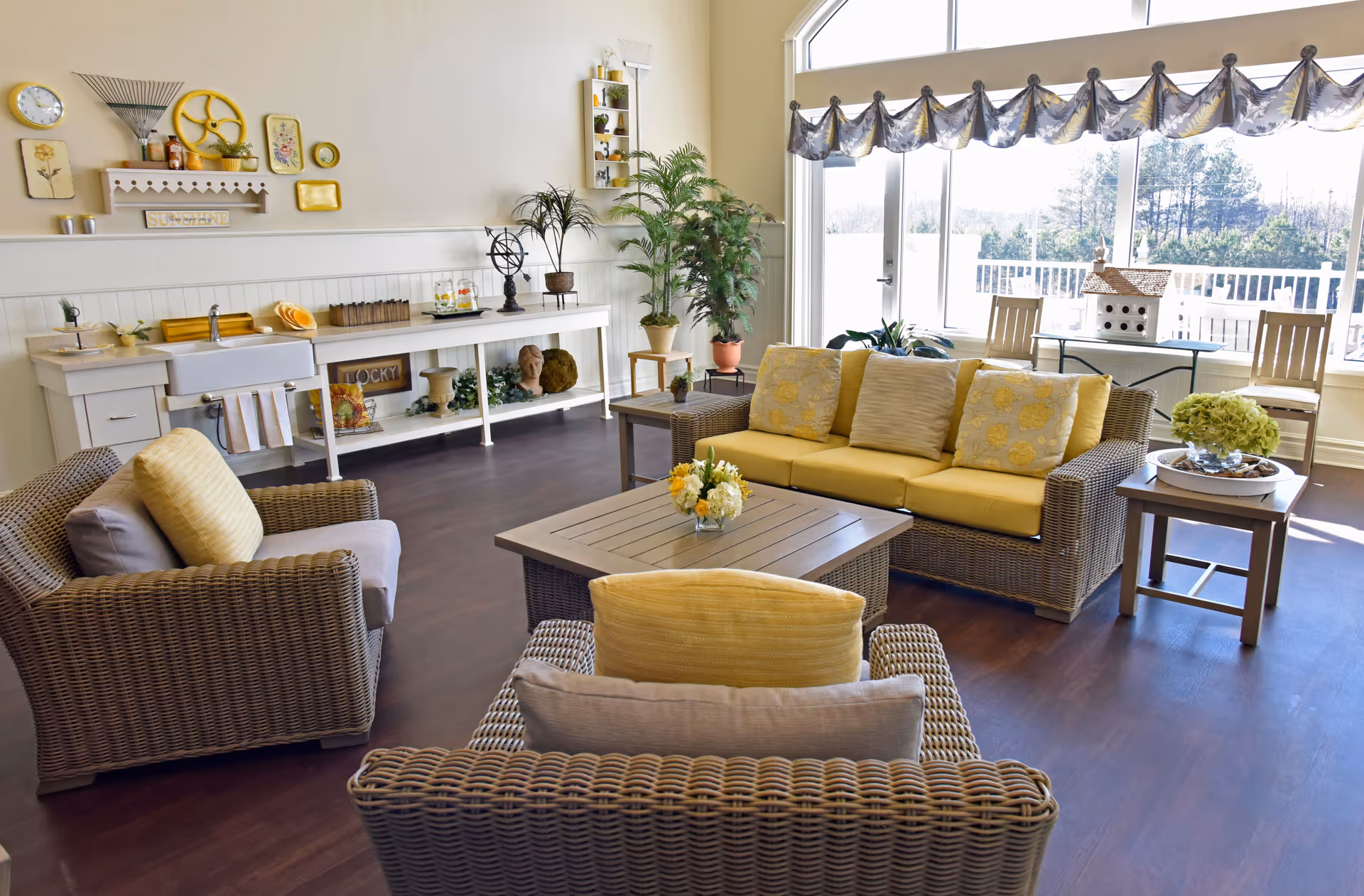 A bright and cozy living room area with wicker furniture including a sofa and two armchairs with yellow and beige cushions. A wooden coffee table with a small flower arrangement sits in the center. Large windows with a decorative valance let in natural light and offer a view of trees outside. The room also features a long white console table against the wall with decorative items, plants, and a small sink. The floor is dark wood, and the overall decor is warm and inviting.