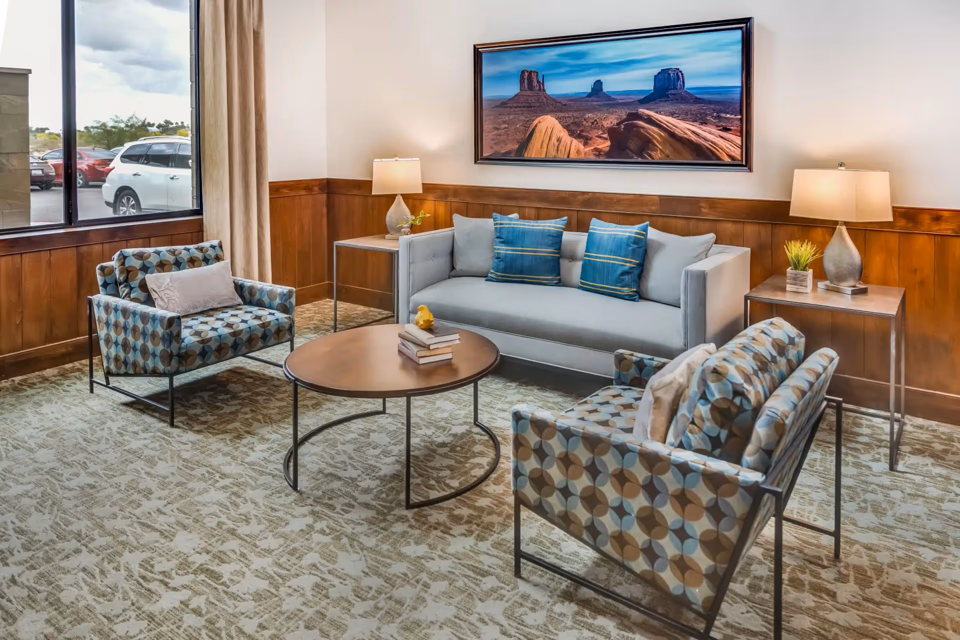 A cozy living room area with a light gray sofa adorned with blue and gray pillows, two patterned armchairs with circular designs, a round wooden coffee table with books and a small decorative item, two side tables each with a lamp and a small plant, wood paneling on the walls, a large window showing parked cars outside, and a framed landscape photograph of a desert scene hanging above the sofa.
