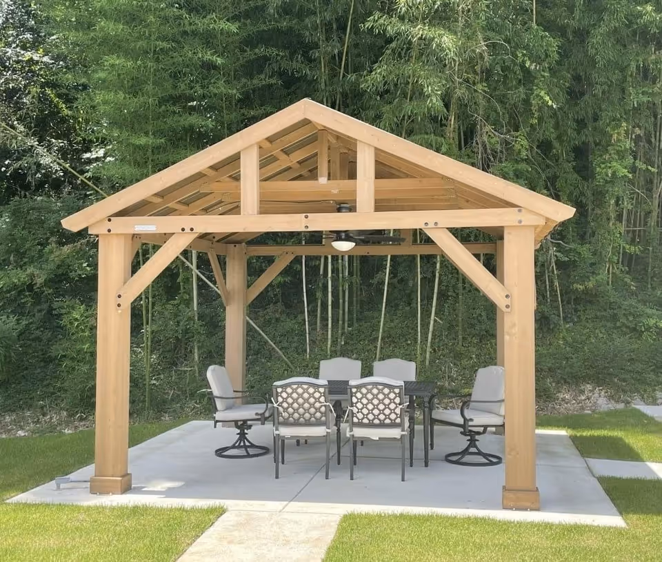 Outdoor wooden gazebo with a peaked roof, containing a black metal table and six cushioned chairs on a concrete slab, surrounded by green grass and tall bamboo trees in the background.