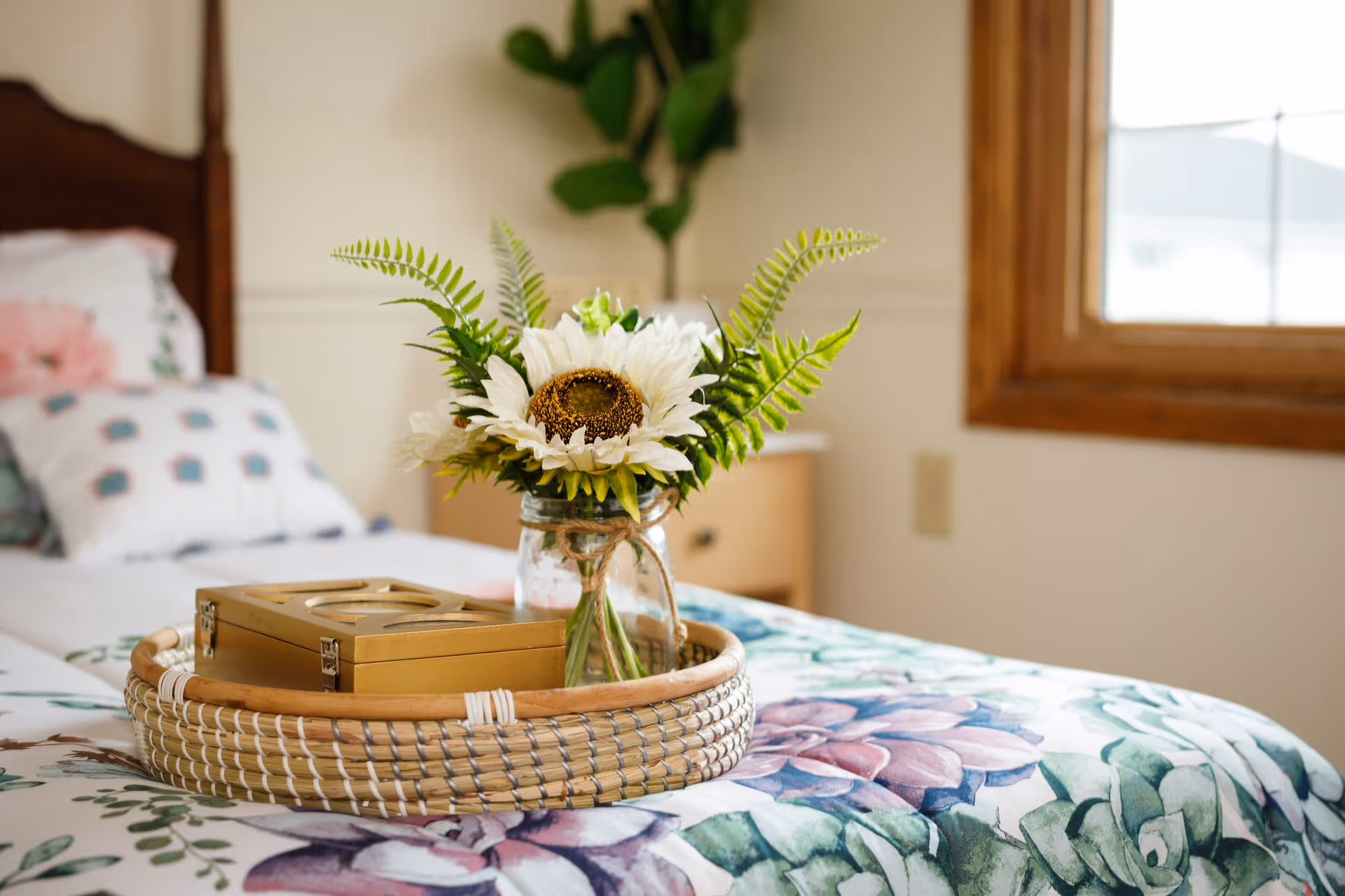 A decorative tray holding a jar of sunflowers and a small box sits on a floral-patterned bed in a bright bedroom.