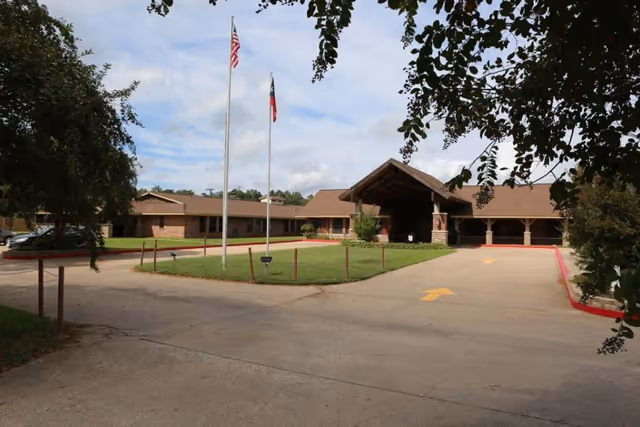 Exterior view of Timberwood Nursing & Rehabilitation Center showing a single-story brick building with a covered entrance, two flagpoles with the American and Texas flags, a driveway with yellow directional arrows, and surrounding greenery including trees and grass.