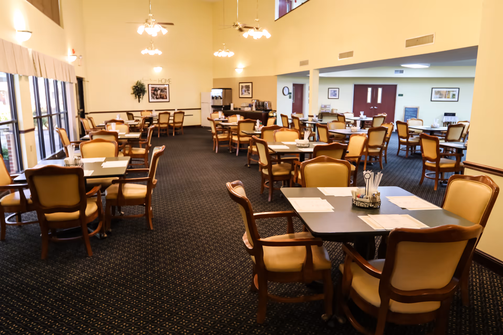 A spacious dining room with multiple tables and chairs arranged neatly. The room has large windows on one side allowing natural light to enter, beige walls, and carpeted flooring. Tables are set with placemats, menus, and condiment holders. Ceiling fans and light fixtures hang from the ceiling.